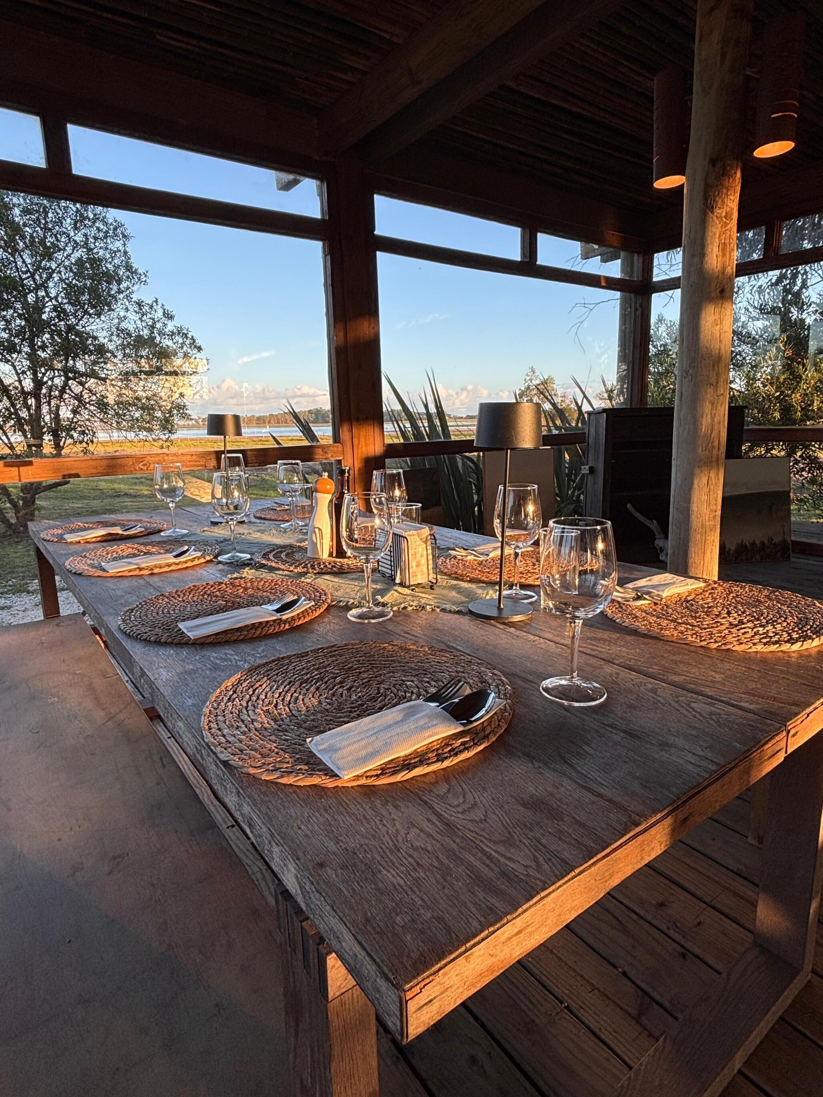 A wooden dining table set with placemats, wine glasses, and cutlery, overlooking a scenic landscape.