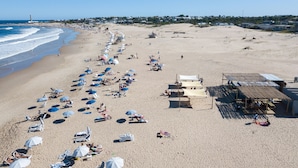 A beach with rows of sun loungers and umbrellas, a lifeguard tower, and a small building, likely a lifeguard station.