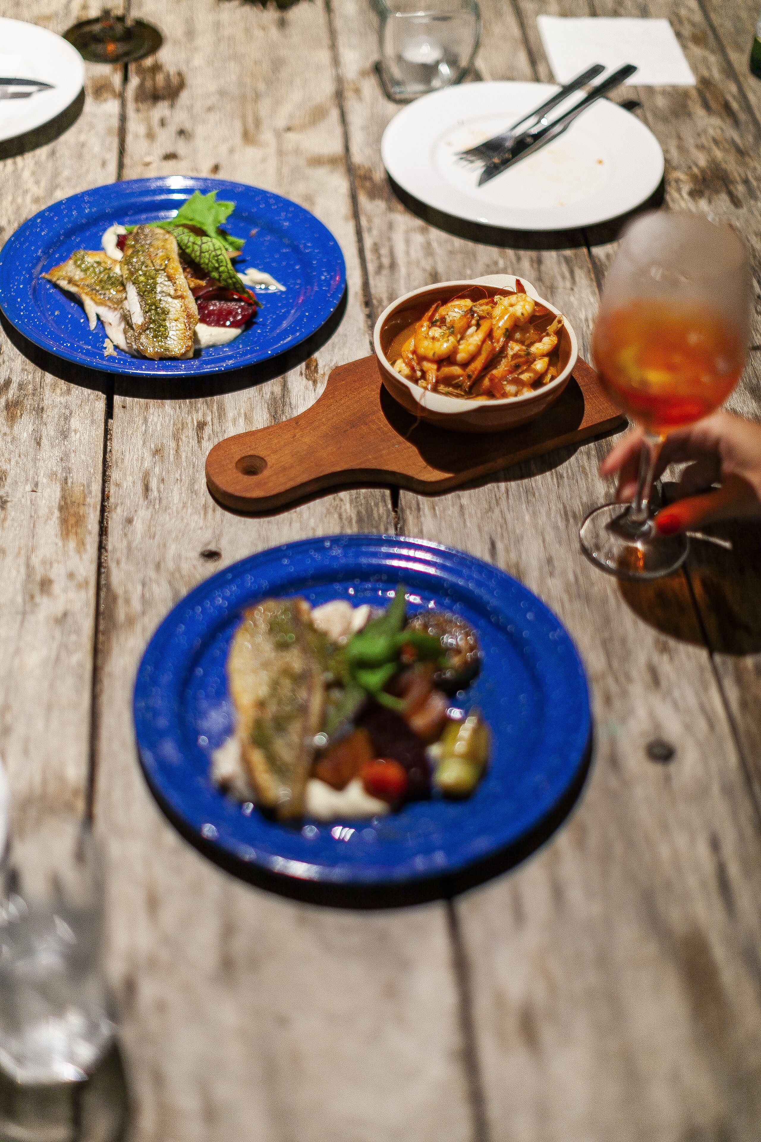 A wooden table with three blue plates of food, a small bowl of shrimp, and a wine glass.