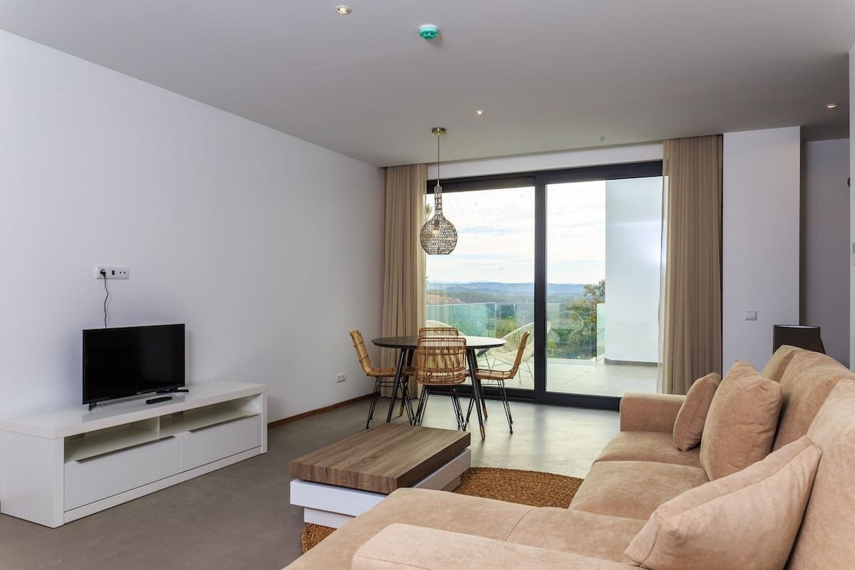 Una sala moderna con un televisor de pantalla plana sobre un mueble blanco, un sofá color beige, una mesa de centro de madera y un comedor con sillas de mimbre y una mesa de vidrio.