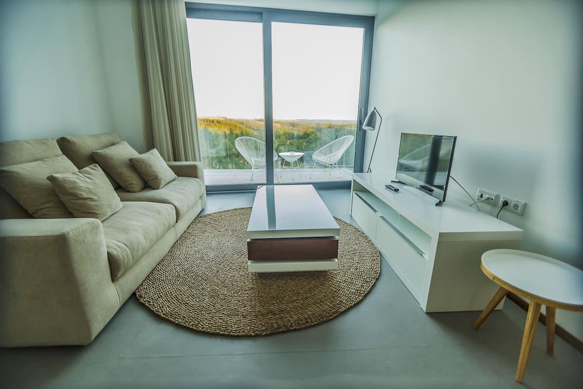 Un salón moderno con un sofá, una mesa de centro, un televisor y vistas amplias a través de grandes ventanales.