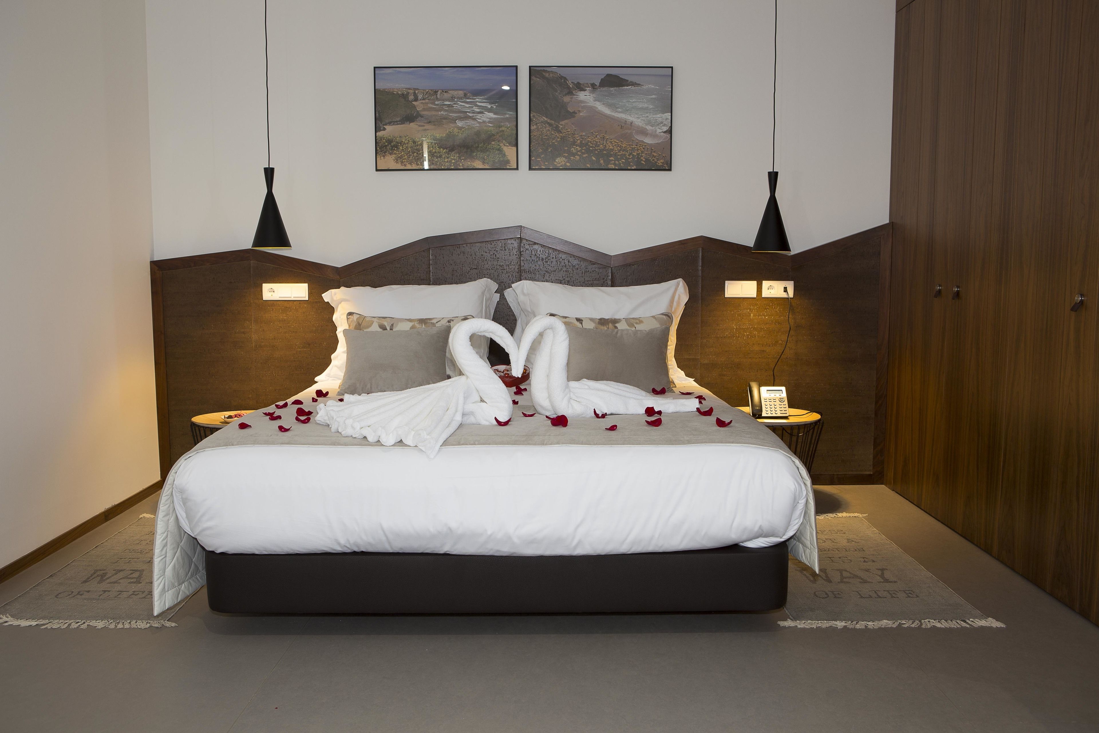 A neatly made bed with white linens and a heart-shaped towel arrangement, two wall-mounted lamps, and two framed pictures above the bed.