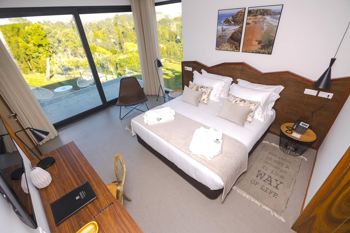 Habitación de hotel moderna con una cama grande, un escritorio y un balcón con vistas a zonas verdes.