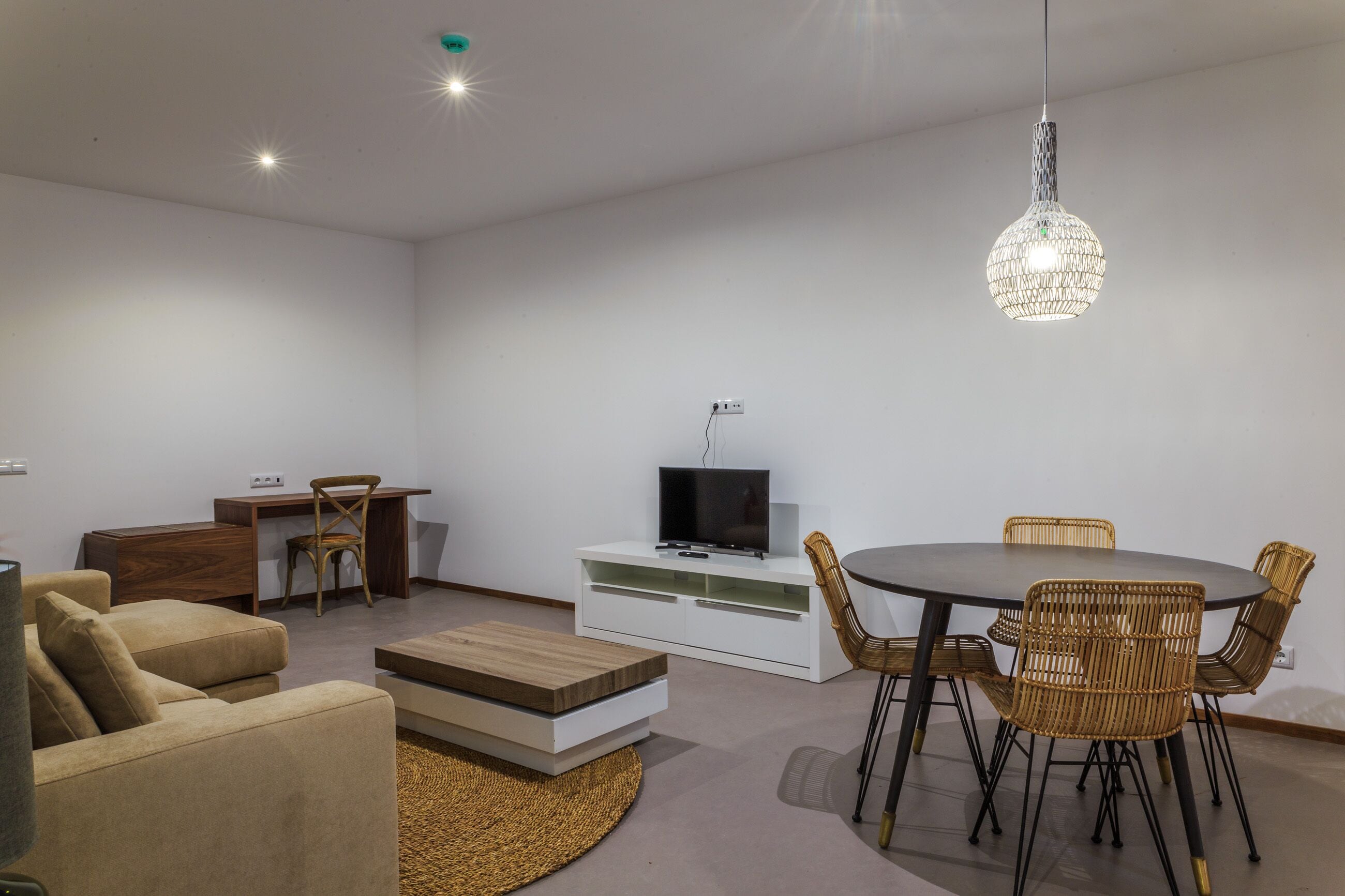 A modern living room with a round dining table, wicker chairs, a flat-screen TV on a white cabinet, and a hanging crystal light fixture.
