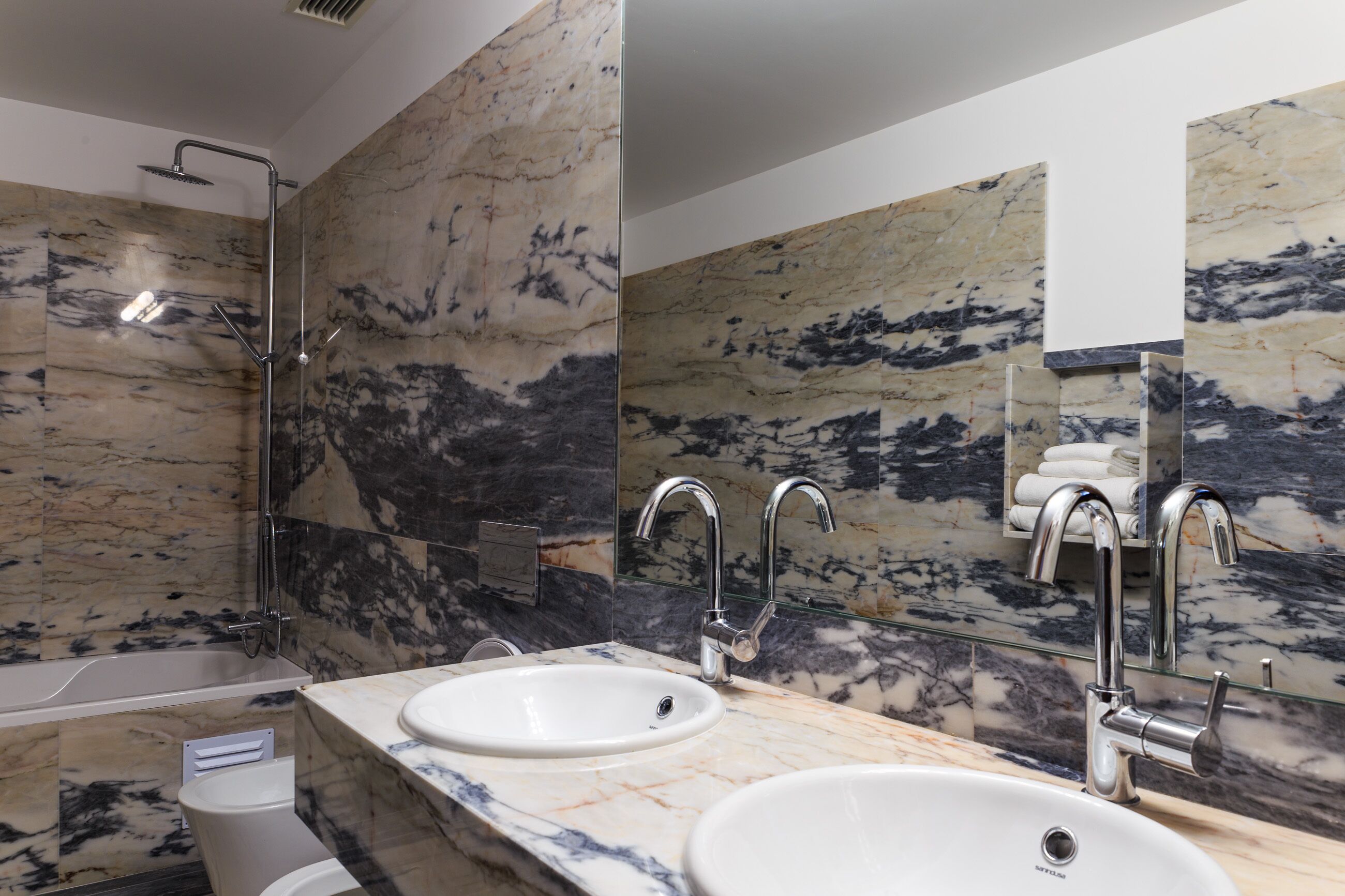 A modern bathroom with marble walls, a bathtub, two sinks, and a glass shower enclosure.