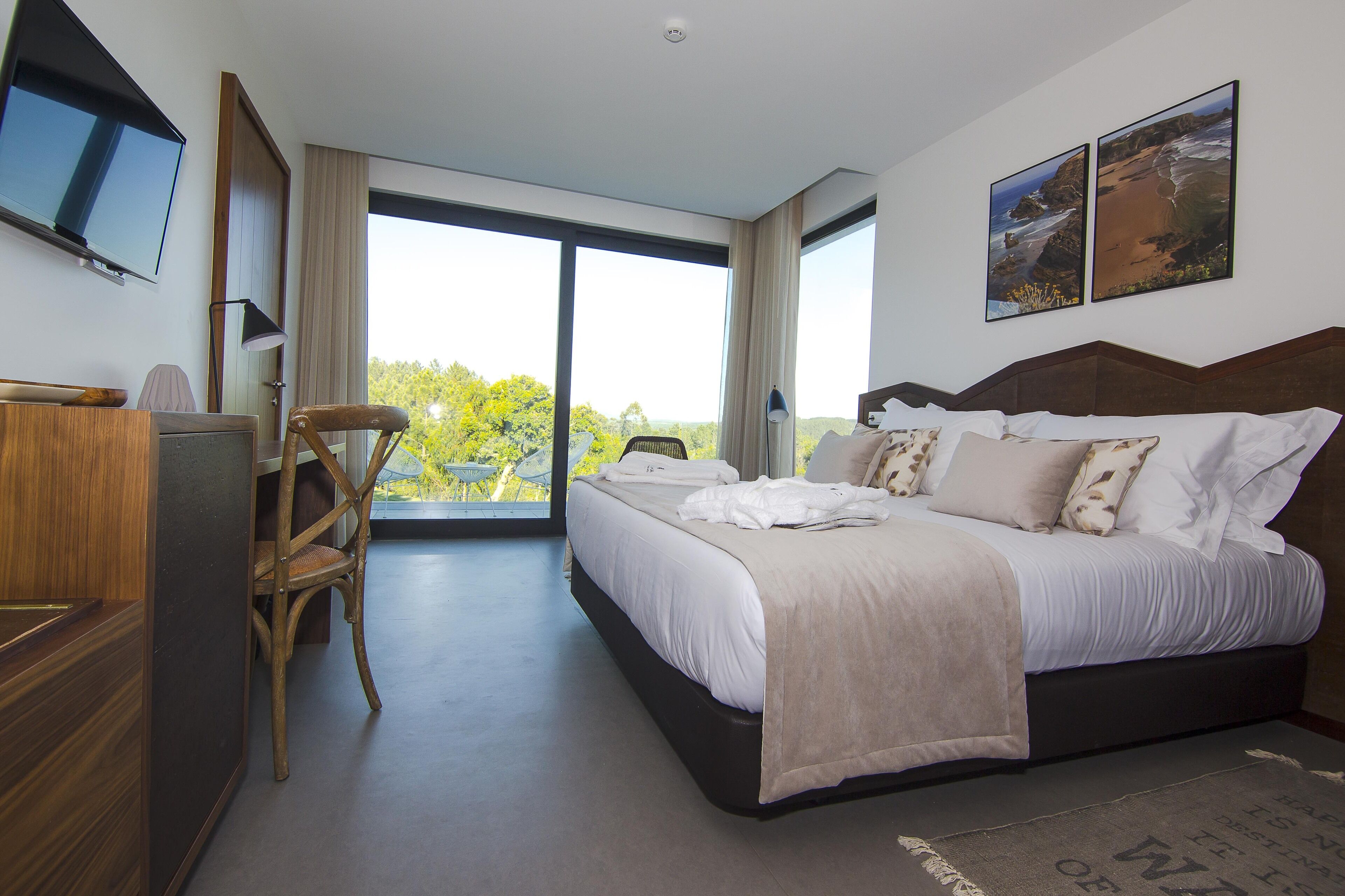 Ein modernes Hotelzimmer mit einem großen Bett, einem Schreibtisch und einem Stuhl. An den Wänden hängen zwei Bilder und durch das Fenster ist ein Blick ins Freie zu sehen.