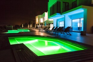 Zona de piscina con sillones de playa y sombrillas, un edificio moderno con ventanas amplias y una piscina bien iluminada con iluminación verde.