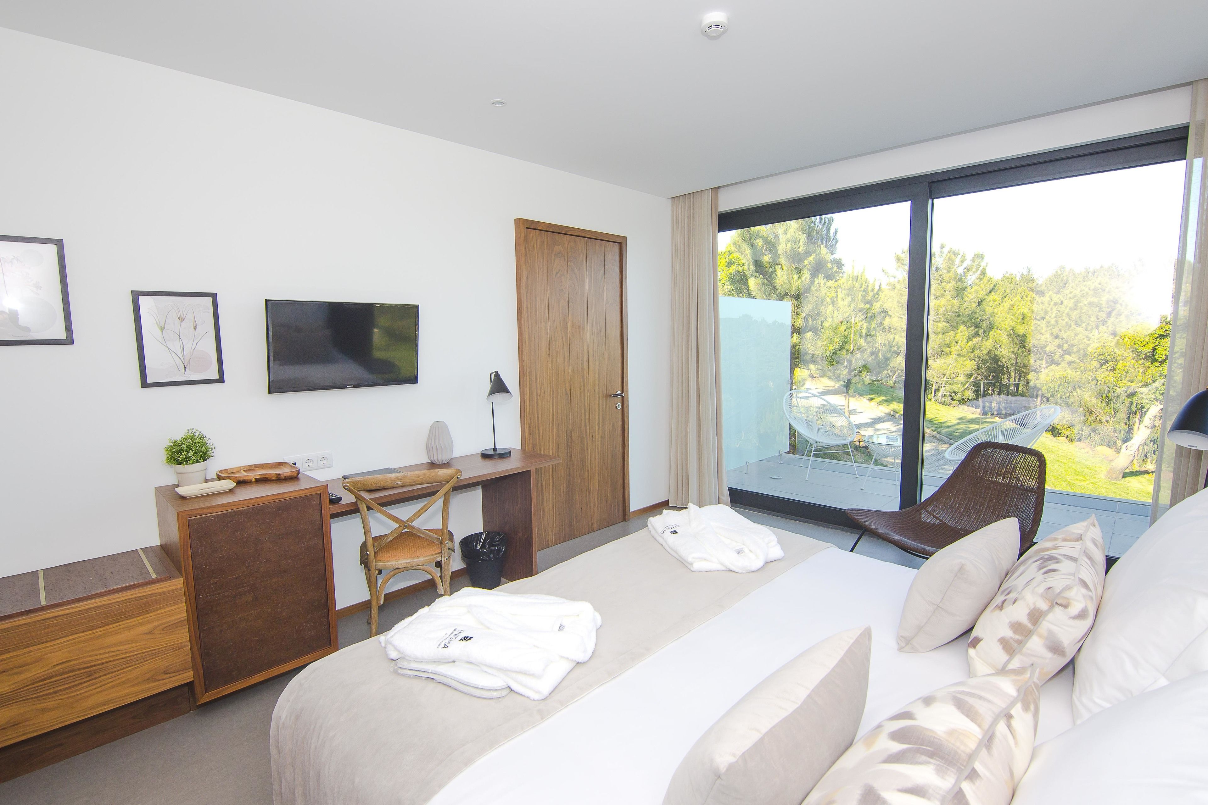 A modern hotel room with a large bed, a desk, and a TV. There is a view of the outdoors through the window.