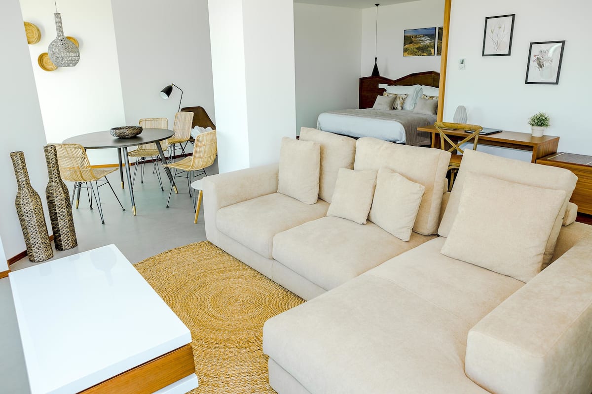 Una sala moderna con un sofá color beige, una mesa de comedor con sillas de mimbre y una cocina al fondo.