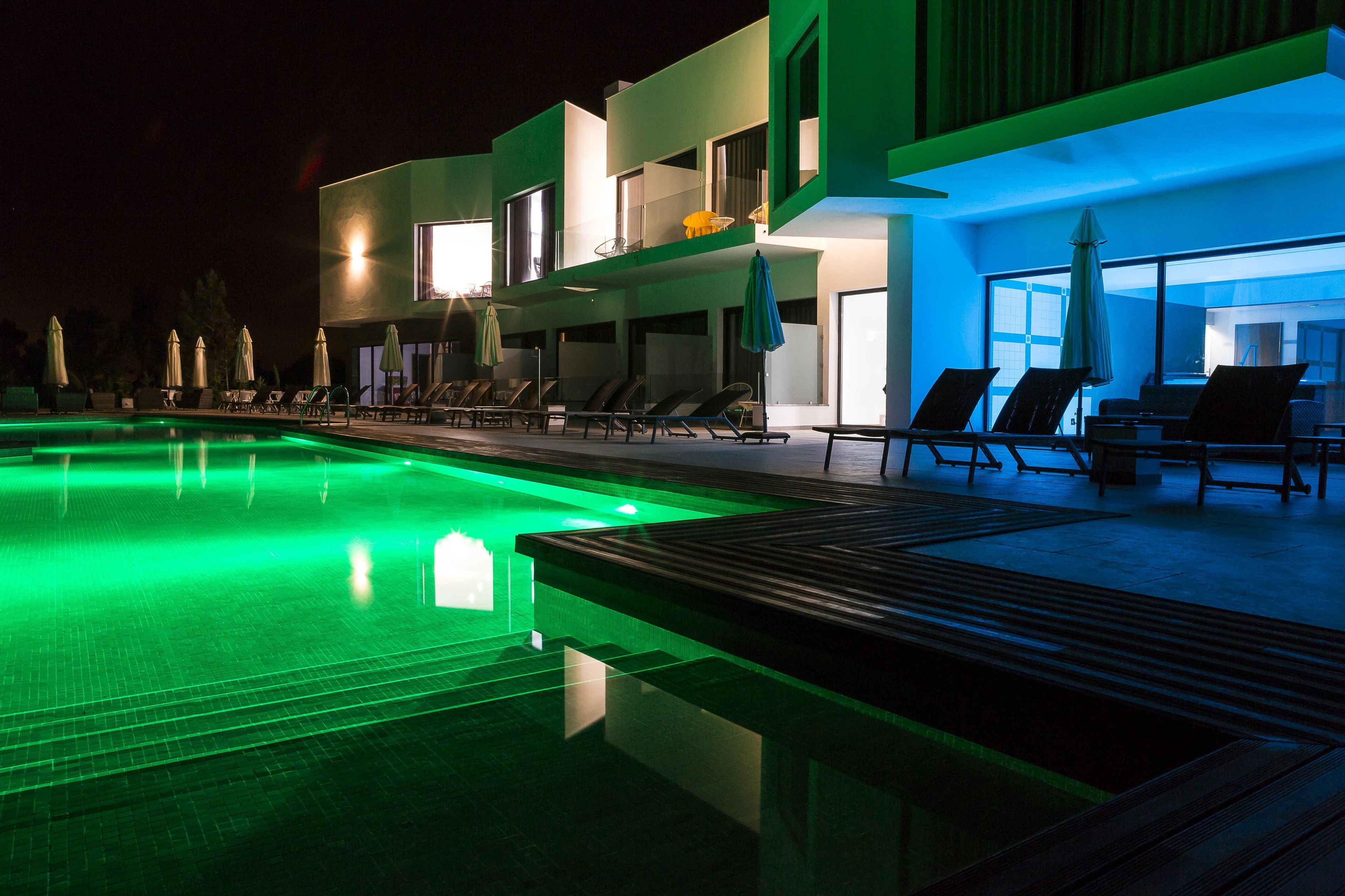 A modern hotel with a swimming pool, sun loungers, and a well-lit exterior at night.