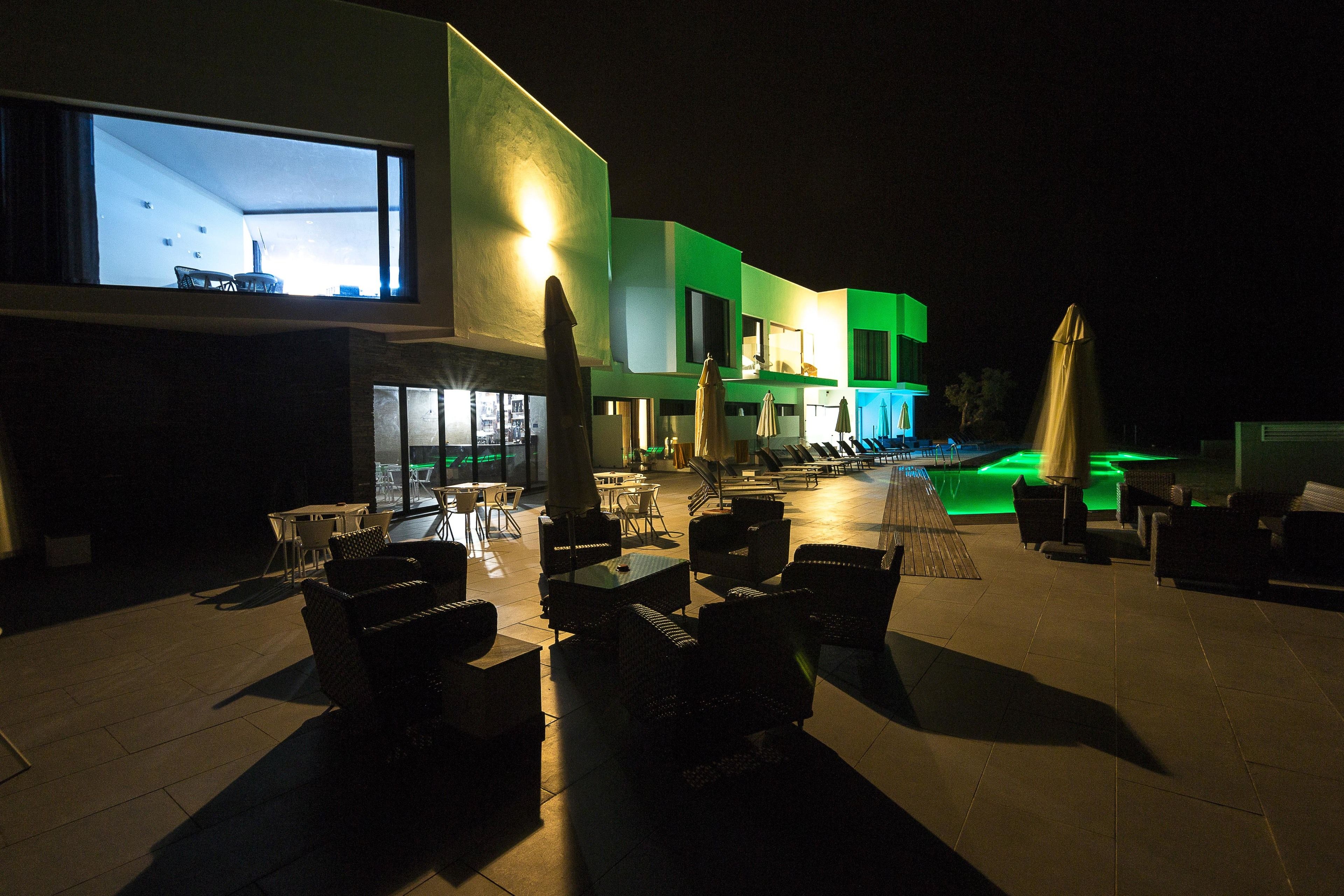 Un espace extérieur aménagé en terrasse, avec des sièges et des parasols, une piscine et un bâtiment dont les fenêtres sont illuminées.