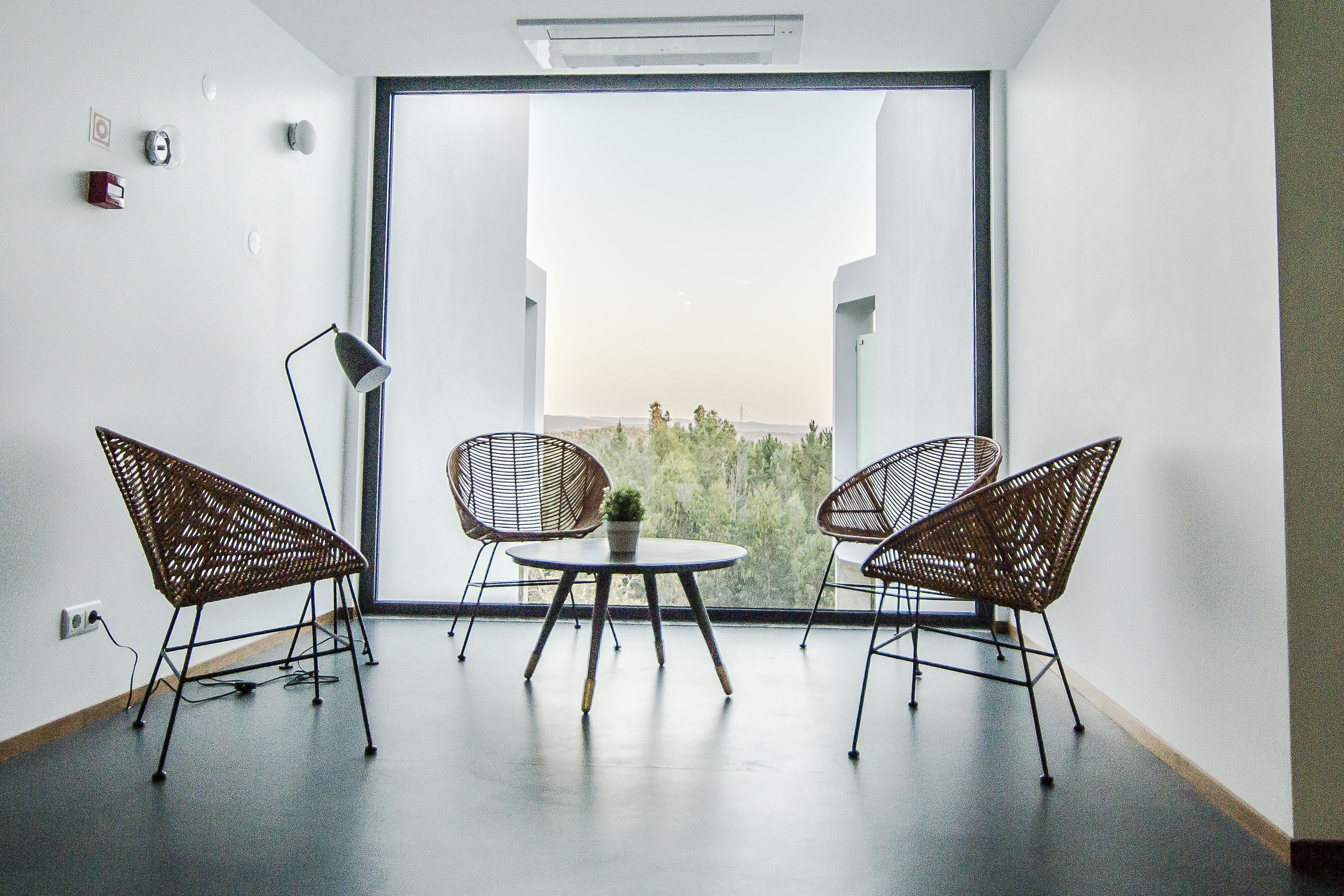 Una habitación moderna con un ventanal grande, dos sillas de mimbre, una mesita y una planta en maceta.