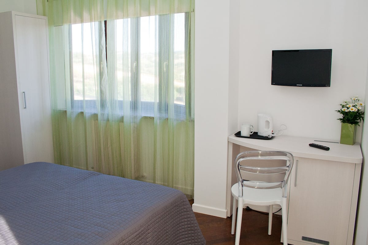 Ein Hotelzimmer mit einem Bett, einem Schreibtisch mit Stuhl, einem Fernseher und einem Fenster mit durchsichtigen Vorhängen.