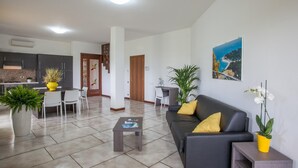 Una sala de estar moderna con sofá, mesa de centro y sillas. Se observa una zona de cocina con armarios y una mesa de comedor.