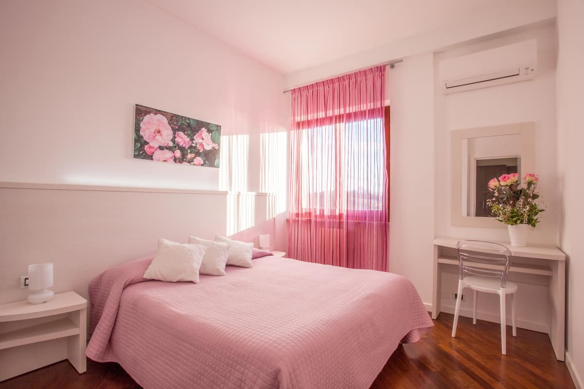 Ein Schlafzimmer mit Bett, Nachttisch, Sessel, einem Fenster mit rosa Vorhängen und einer Wand-Klimaanlage.