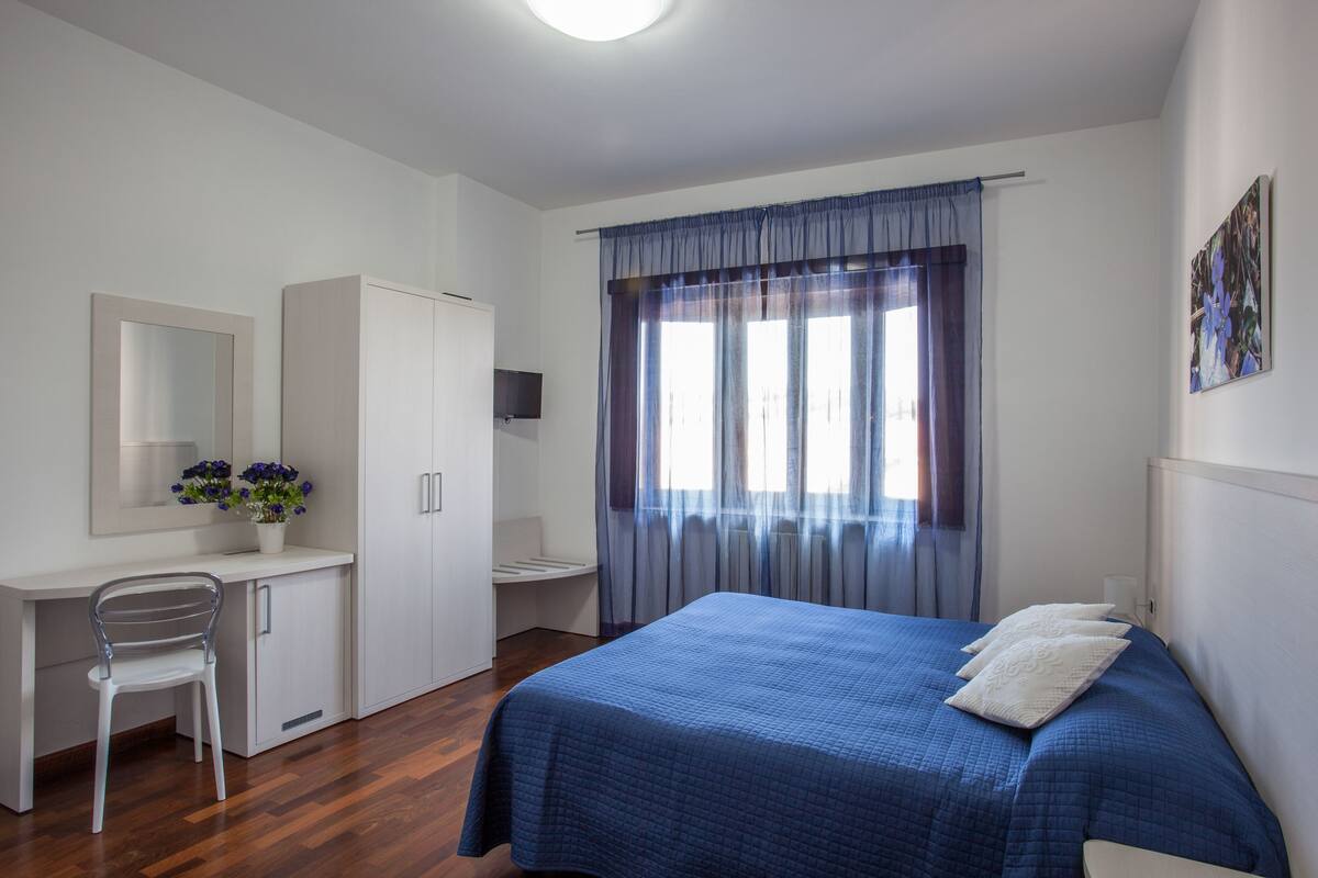 Ein Schlafzimmer mit einem Bett, einem Schreibtisch, einem Stuhl, einem Fernseher und einem Fenster mit Vorhängen.