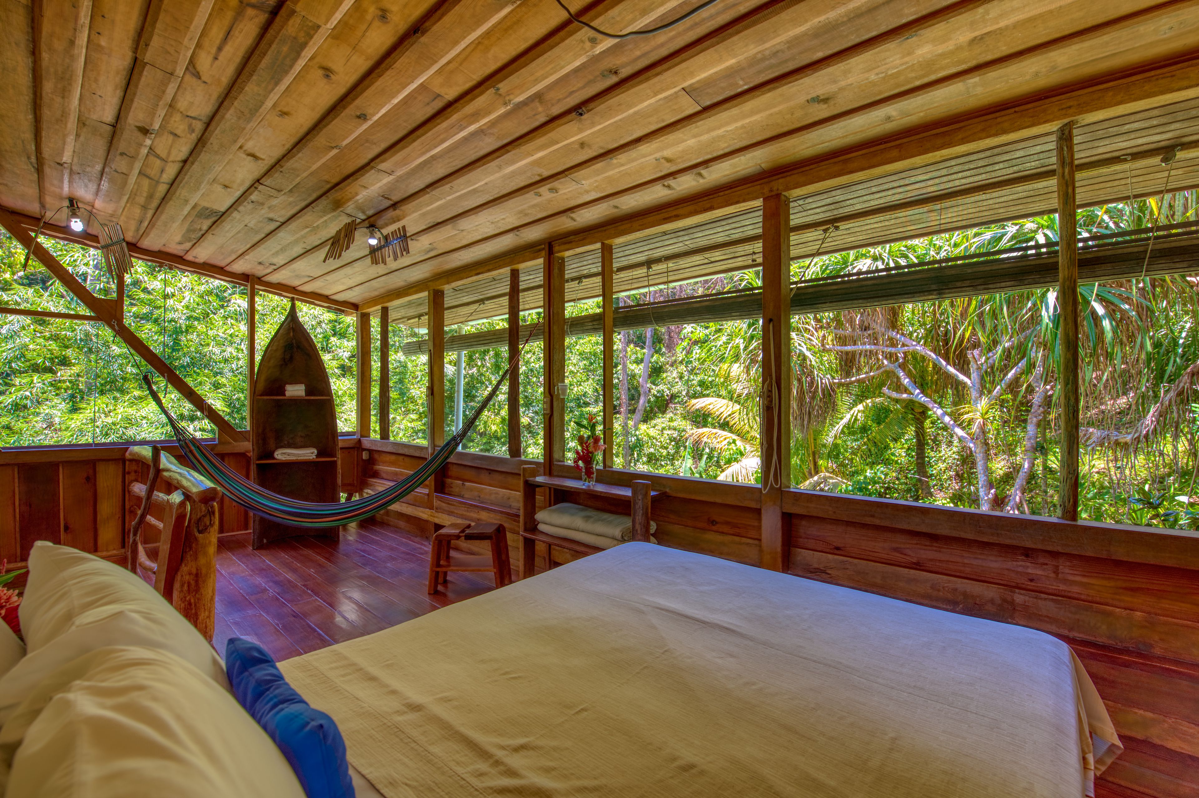 A wooden room with a bed, a hammock, and large windows overlooking a lush garden.