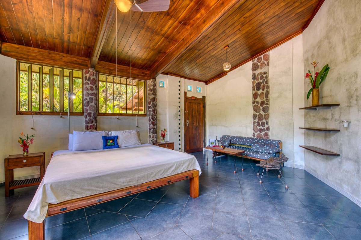 A spacious room with a large bed, a wooden ceiling, stone pillars, a stone wall, and a view of greenery through large windows.