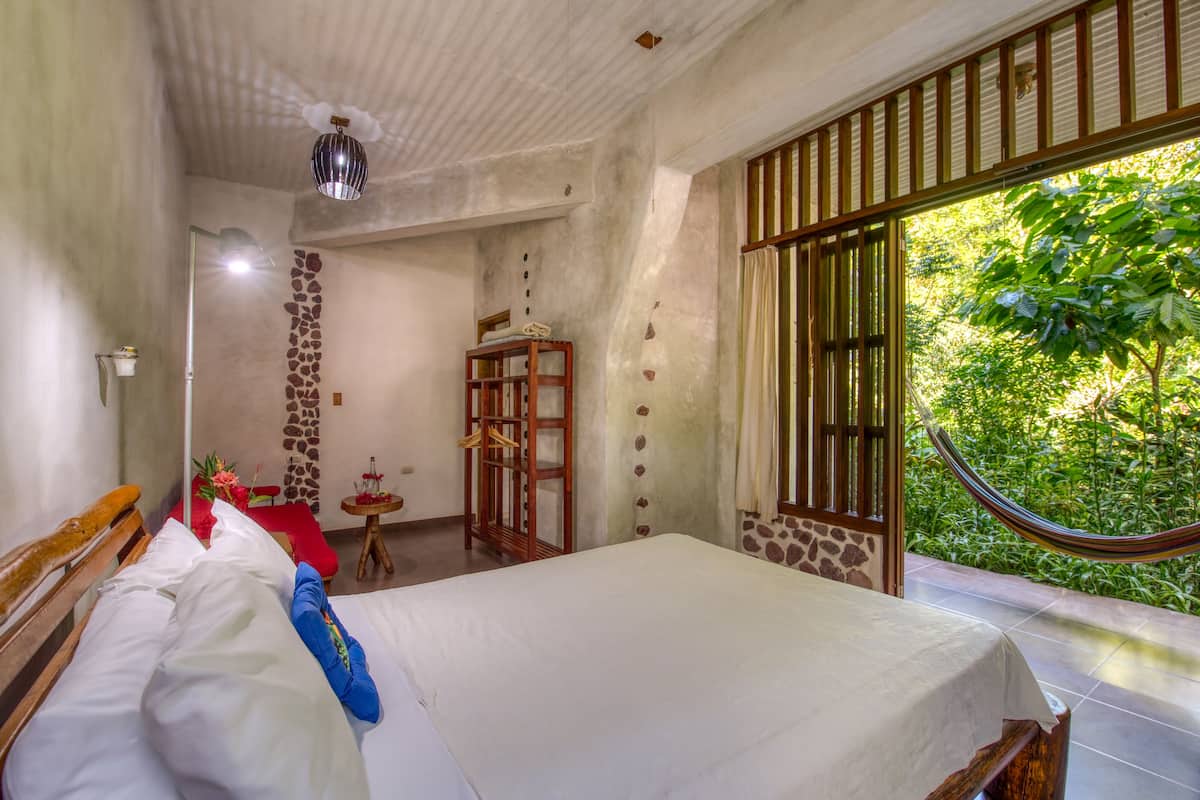 A bedroom with a bed, a wooden wardrobe, a small table with a lamp, and a view of greenery outside.