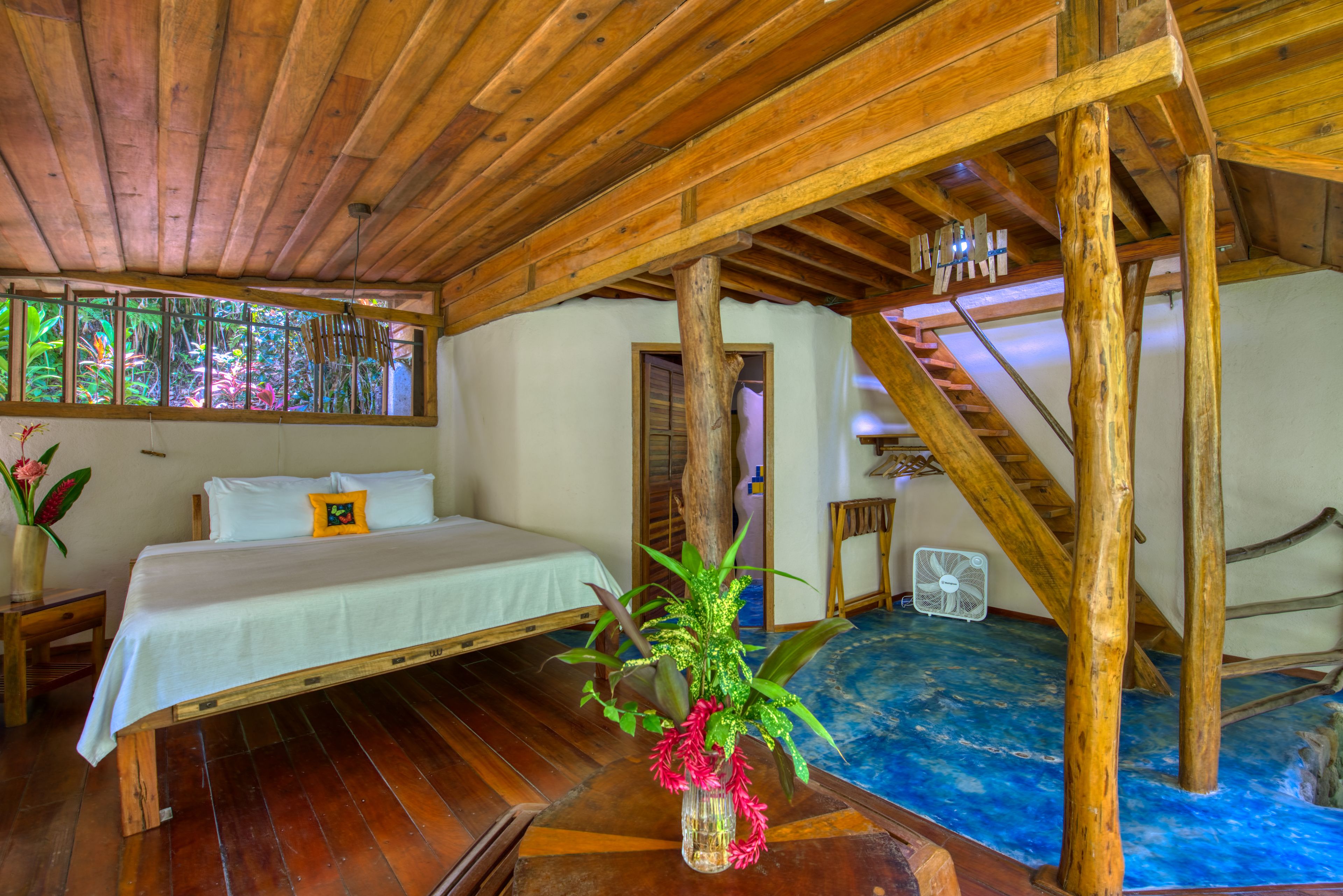 A wooden room with a bed, a fan, and a window with a view of greenery.