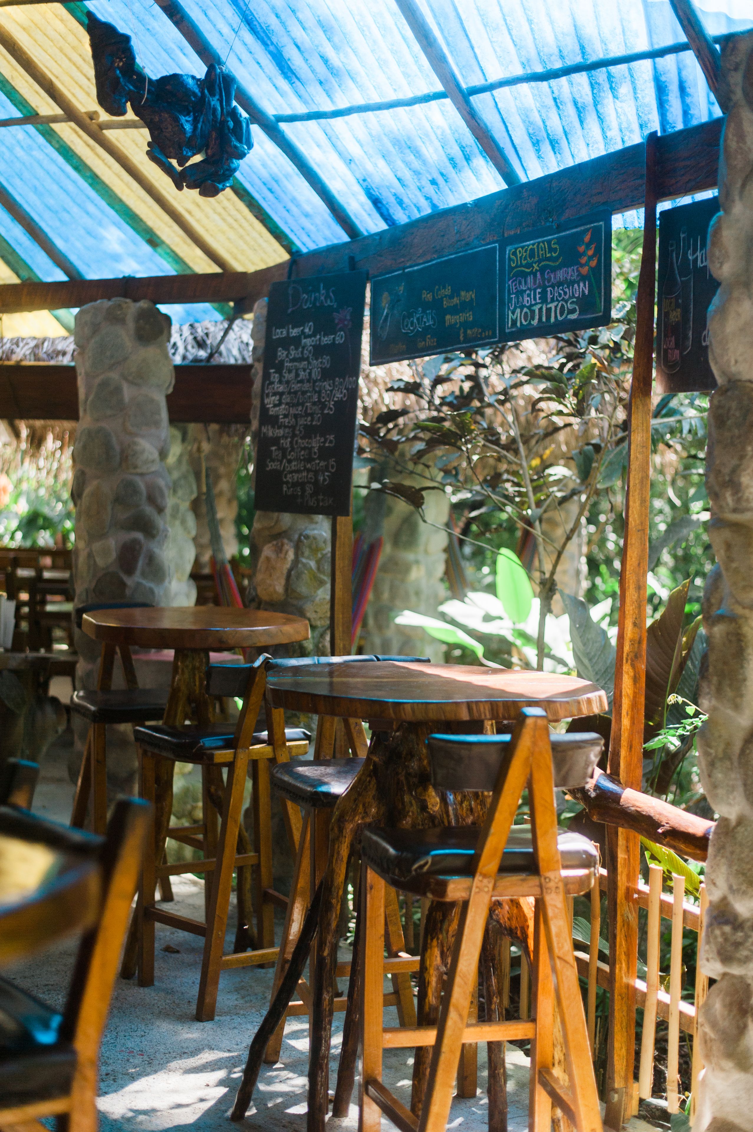 A rustic outdoor café with wooden tables and chairs, a stone pillar, and a menu board listing specials.