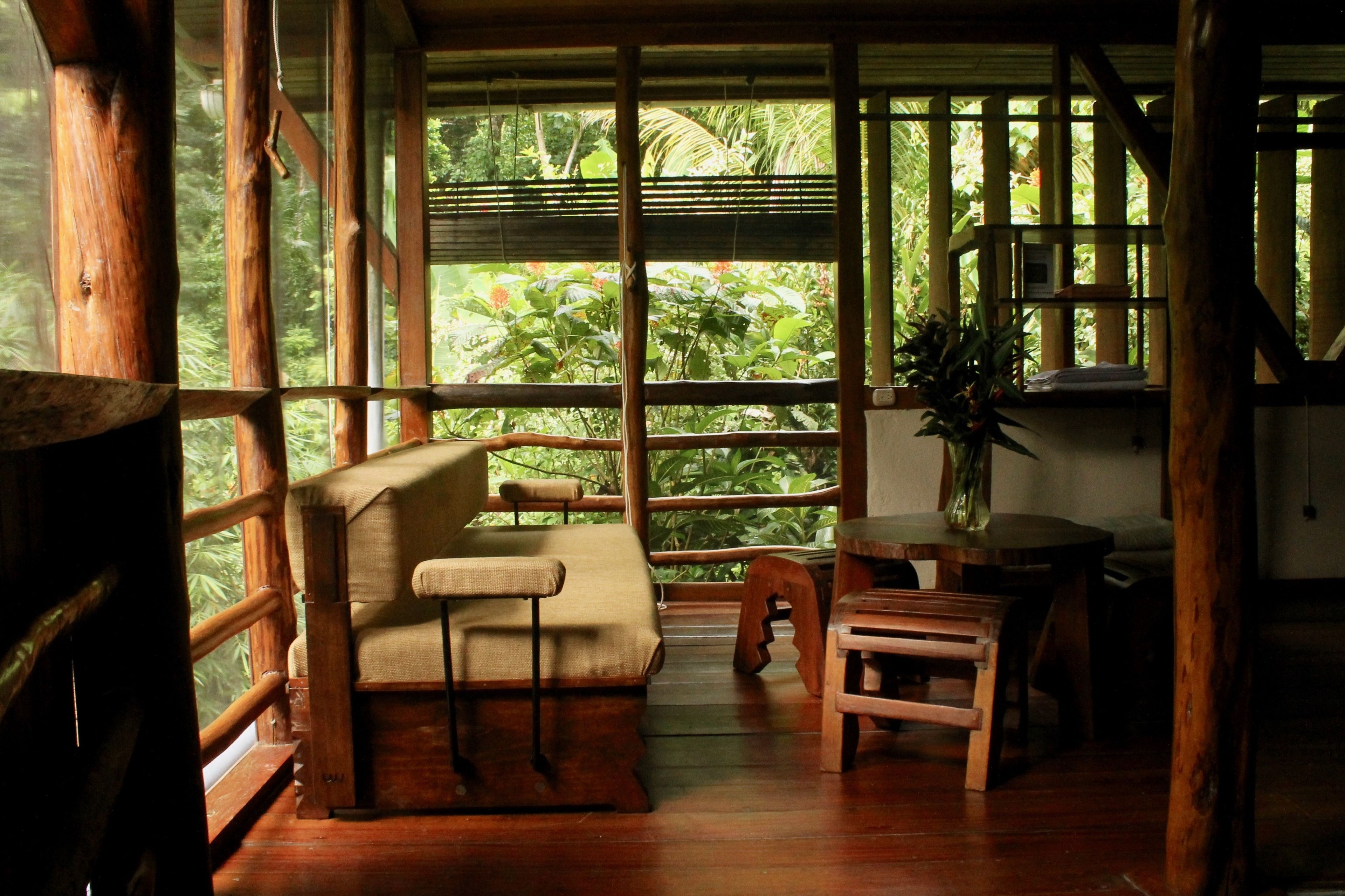 A wooden room with a sofa, a small table, and a chair. Large windows allow natural light and views of greenery outside.