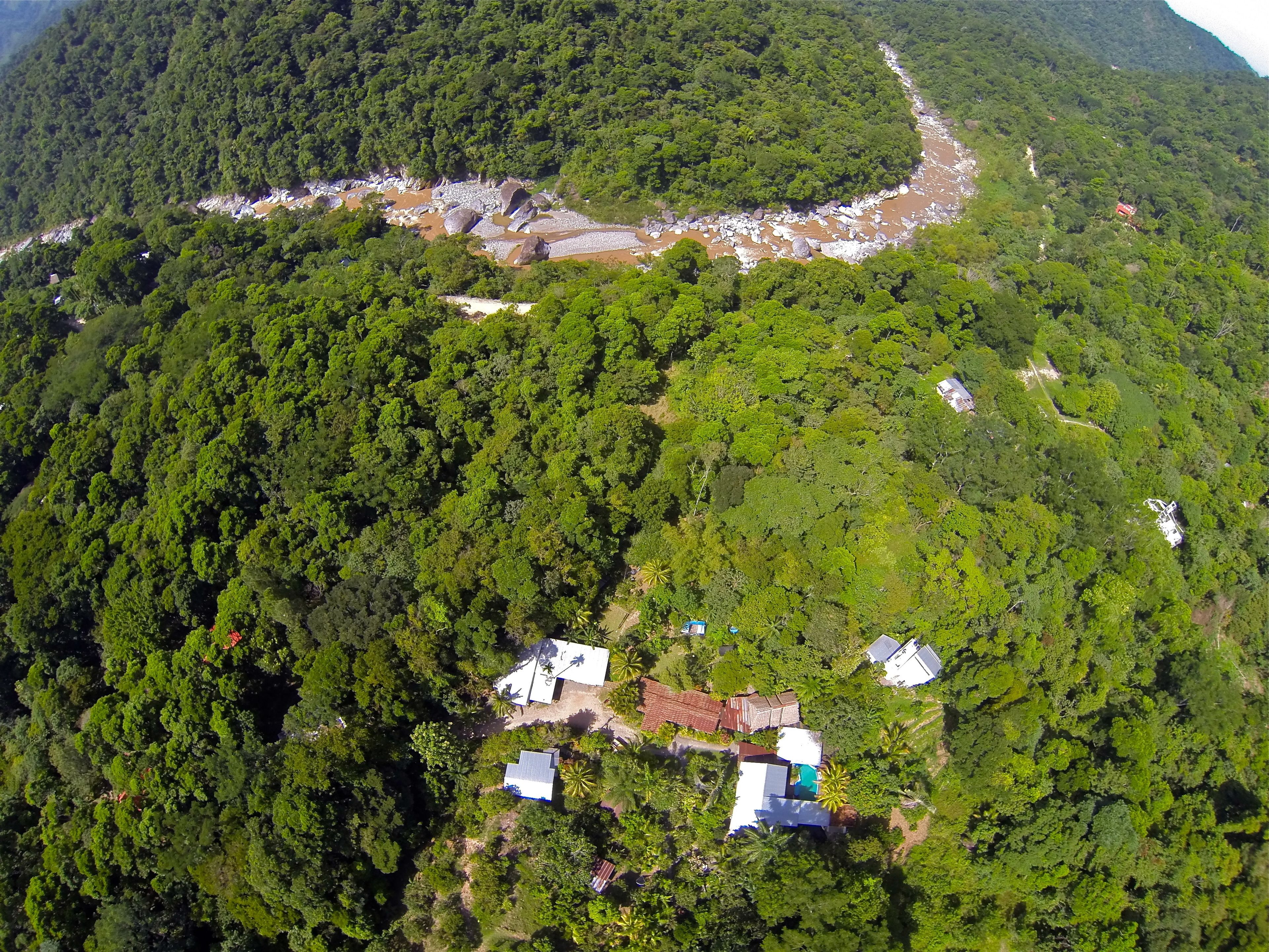 An aerial view of a forested area with several buildings, including houses and a structure with a red roof, surrounded by dense greenery.