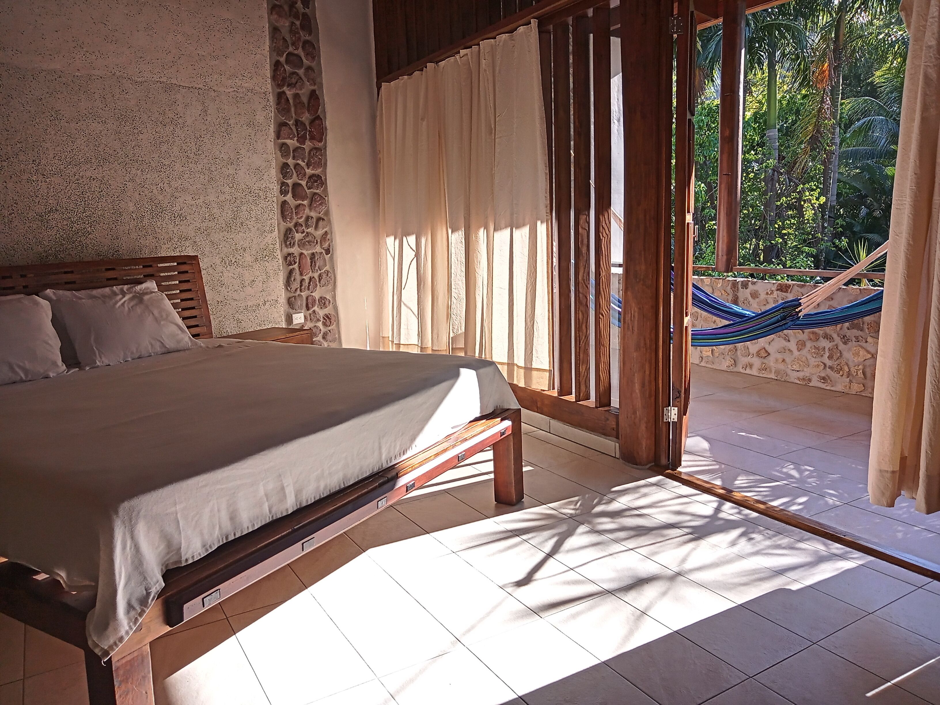 A bedroom with a bed, wooden headboard, stone wall, and a view of a hammock and greenery outside.