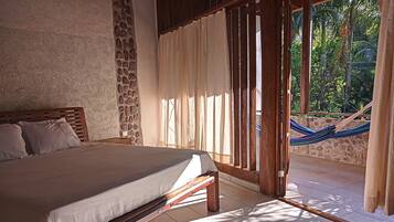 A bedroom with a bed, wooden headboard, stone wall, and a view of a hammock and greenery outside.