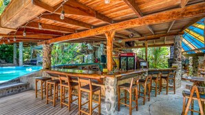 A wooden bar area with stone pillars, a pool, and tropical plants.