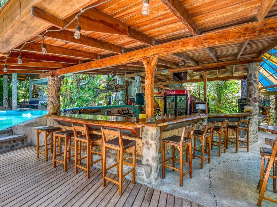 A wooden bar area with stone pillars, a pool, and tropical plants.
