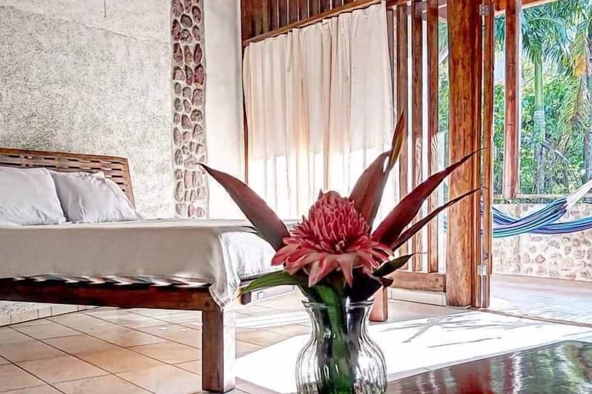 A room with a bed, a table with a vase of flowers, and a view of greenery outside.