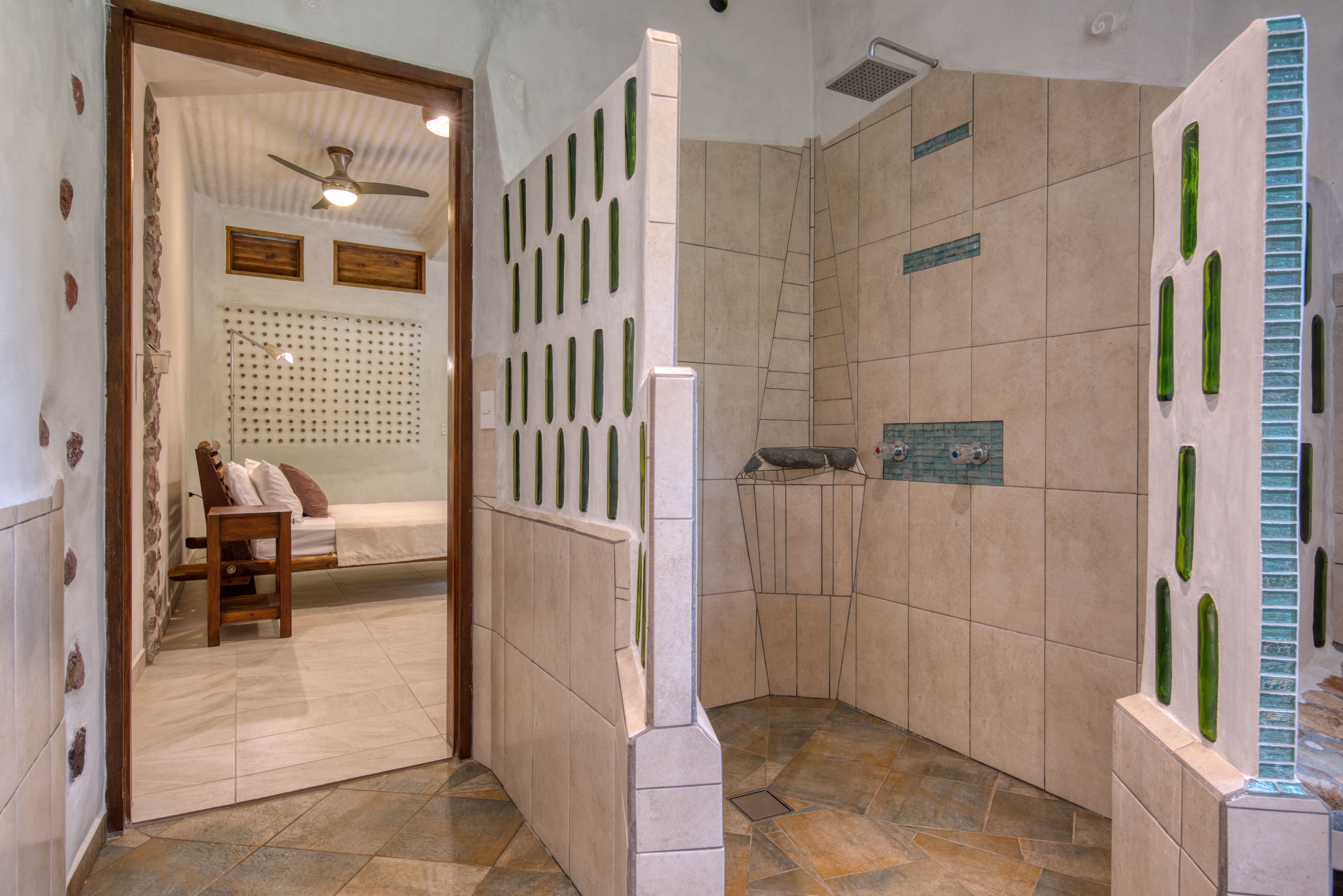 A bathroom with a shower, a toilet, and a sink. There is a bed with a wooden headboard and a pillow in the adjacent room.
