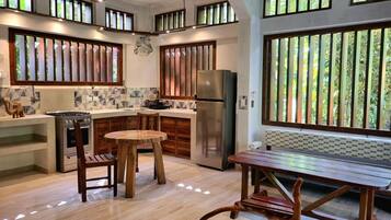A modern kitchen with a central island, wooden chairs, a dining table, and large windows with vertical blinds.