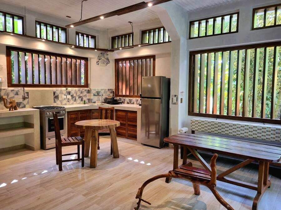 A modern kitchen with a central island, wooden chairs, a dining table, and large windows with vertical blinds.