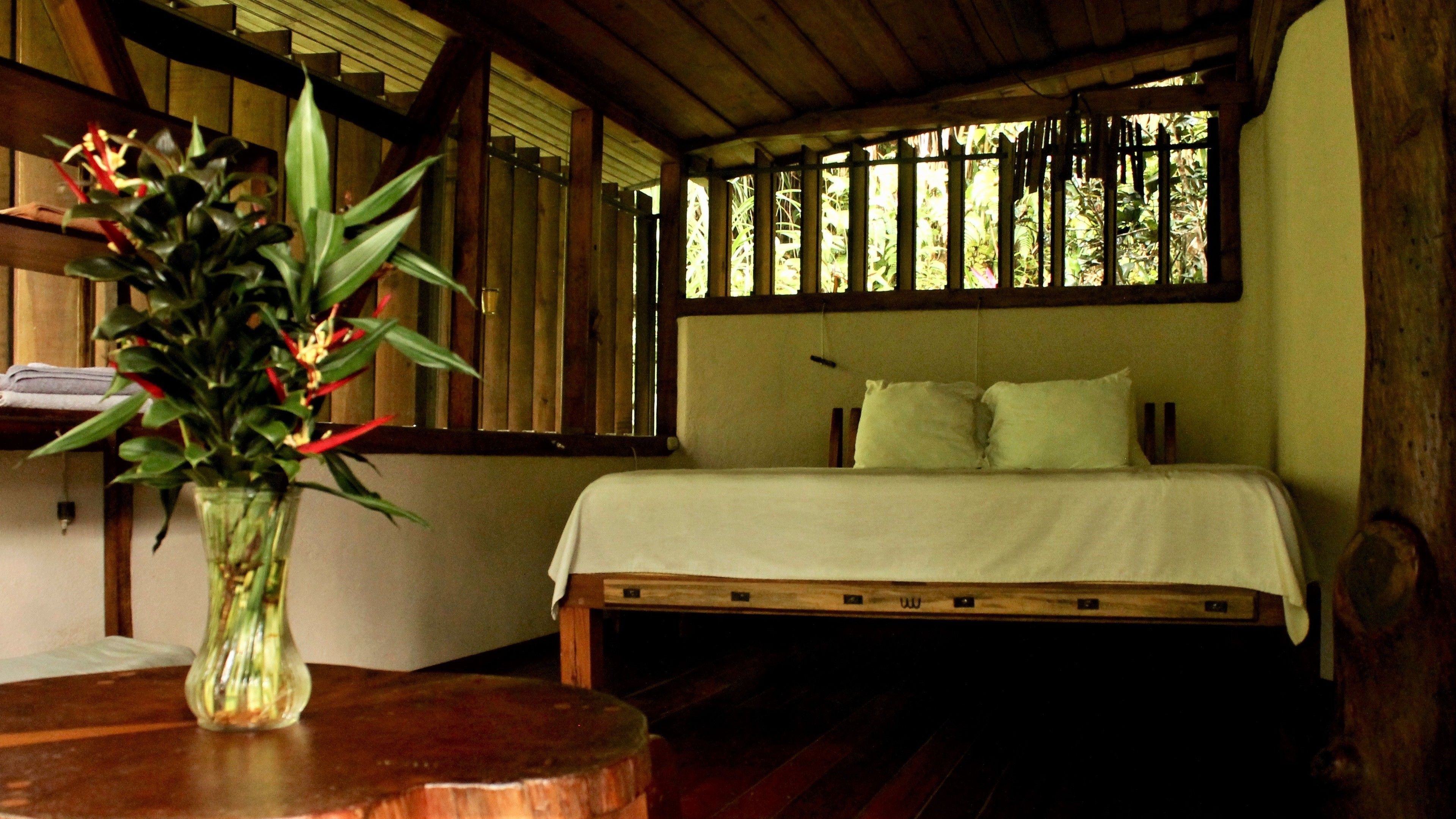 A room with a bed, a wooden headboard, a window with vertical blinds, and a vase with flowers on a wooden table.