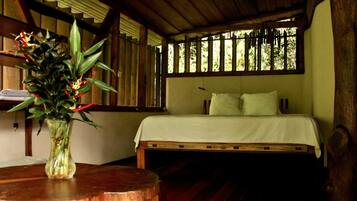 A room with a bed, a wooden headboard, a window with vertical blinds, and a vase with flowers on a wooden table.