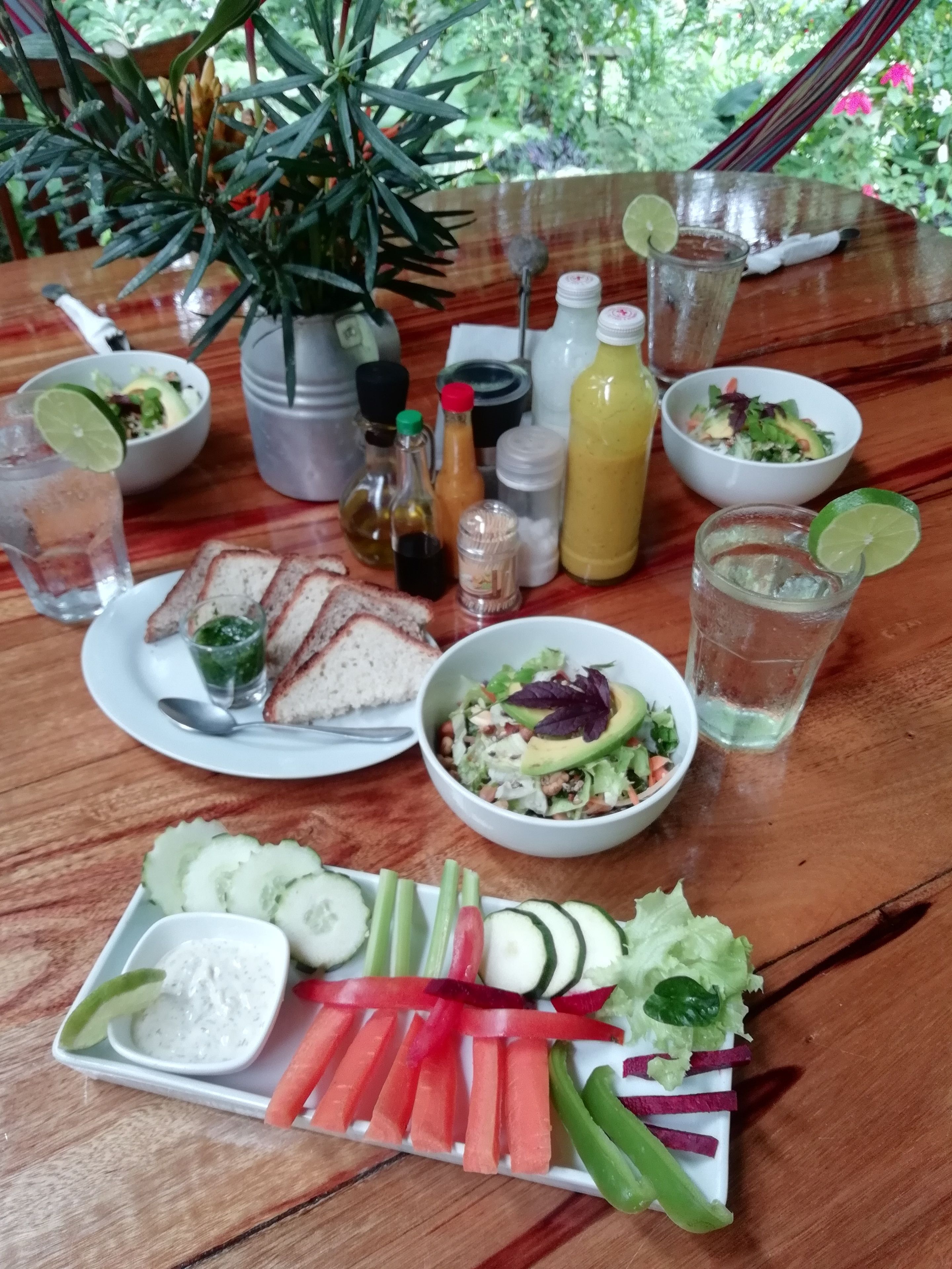 A wooden table with a variety of food items, including sliced cucumbers, carrots, and a salad with avocado and lettuce. There are also condiments and drinks on the table.