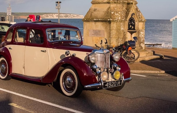 Une voiture ancienne est garée au bord de la mer, avec un monument en arrière-plan.