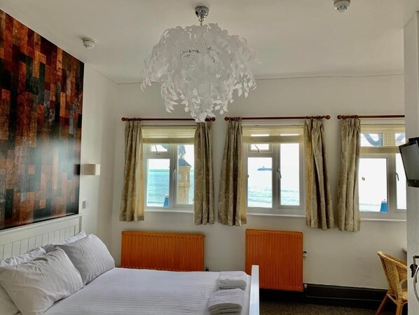 A hotel room with a bed, two windows with curtains, a radiator, a wall-mounted light fixture, and a view of the sea. - The Waterfront Inn (Shanklin)