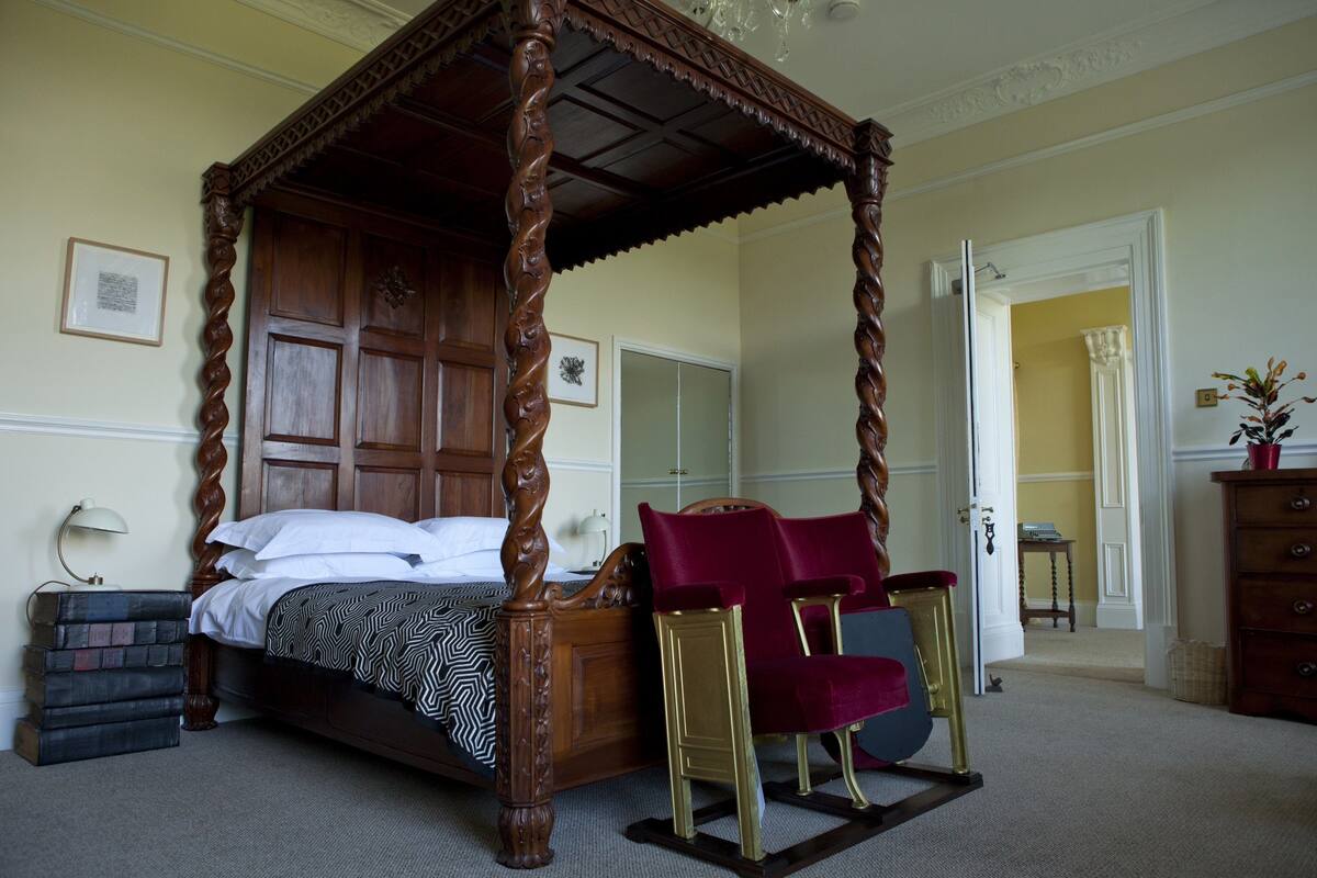A four-poster bed with a wooden frame and a patterned bedspread, a red upholstered chair, a wooden dresser, and a nightstand with books.
