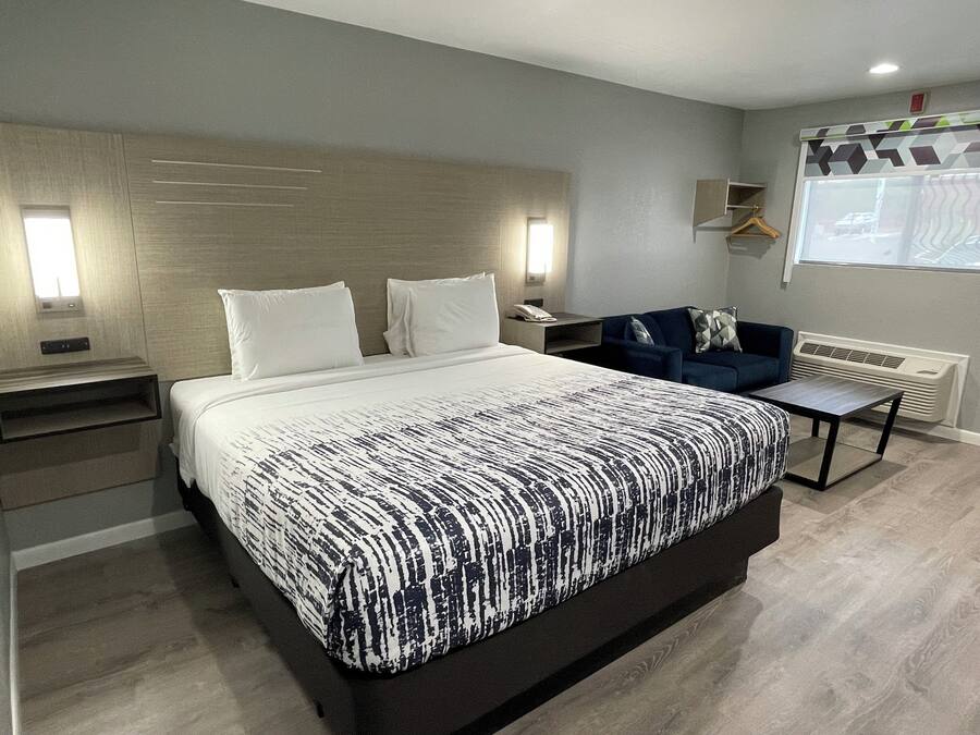 A modern hotel room with a large bed, bedside tables, a sofa, and a window with blinds.
