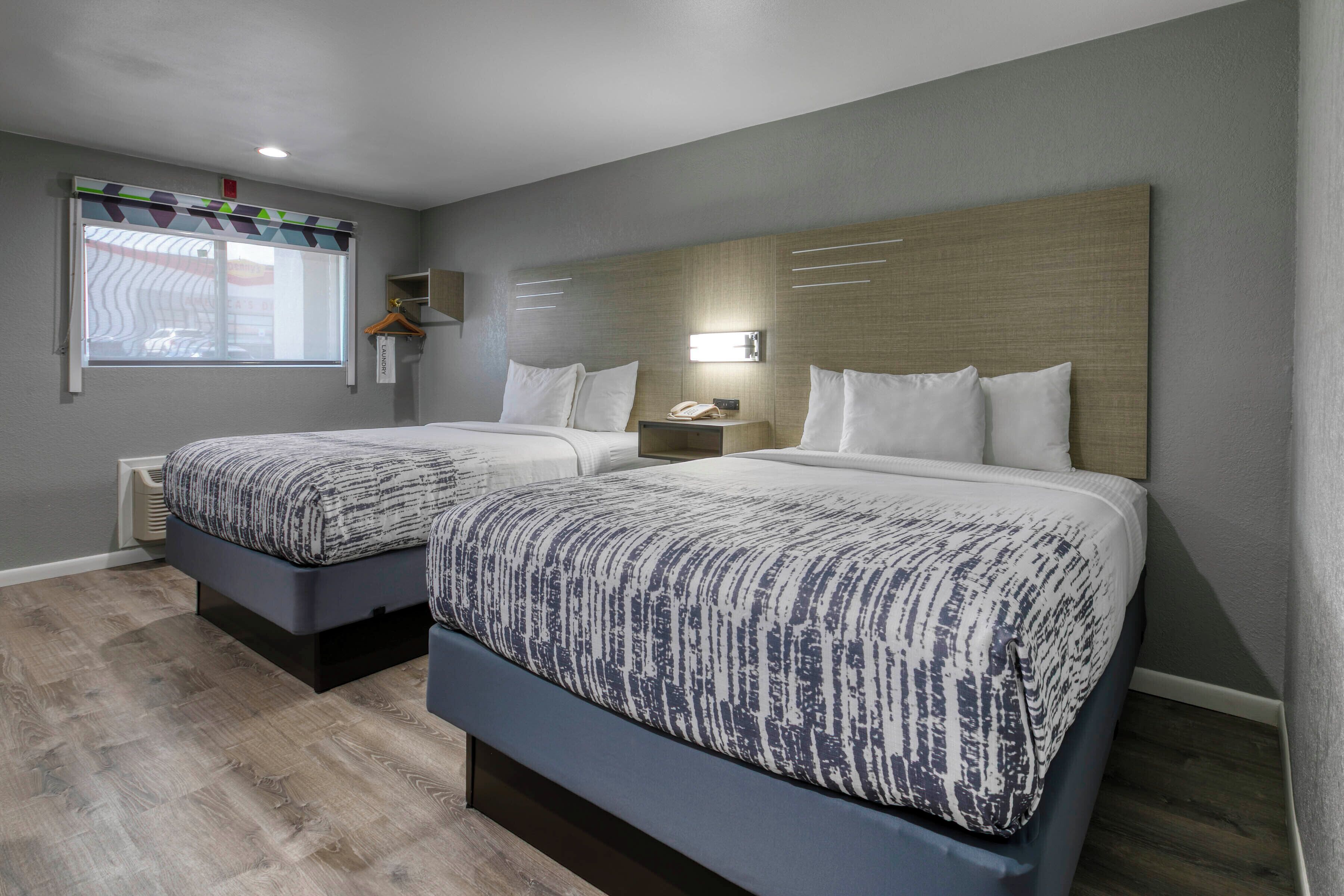 A hotel room with two beds, a window with blinds, a bedside table with a lamp, and a wall-mounted shelf.