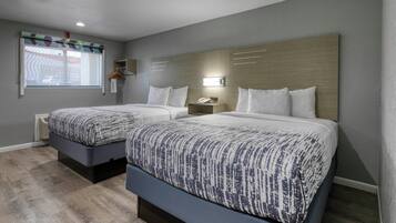 A hotel room with two beds, a window with blinds, a bedside table with a lamp, and a wall-mounted shelf.