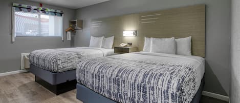 A hotel room with two beds, a window with blinds, a bedside table with a lamp, and a wall-mounted shelf.