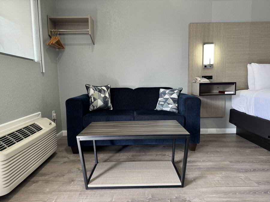 A modern hotel room with a dark blue sofa, two patterned pillows, a small table, a heater, a bed with white linens, and a wooden headboard.