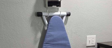 A wall-mounted ironing board with an iron, a power outlet, and a trash bin.