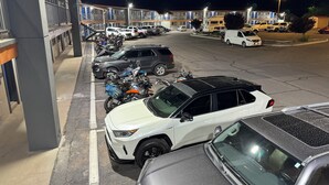 Un pàrquing amb diversos vehicles, incloent cotxes i motocicletes, davant d'un edifici de molts pisos i balcons.