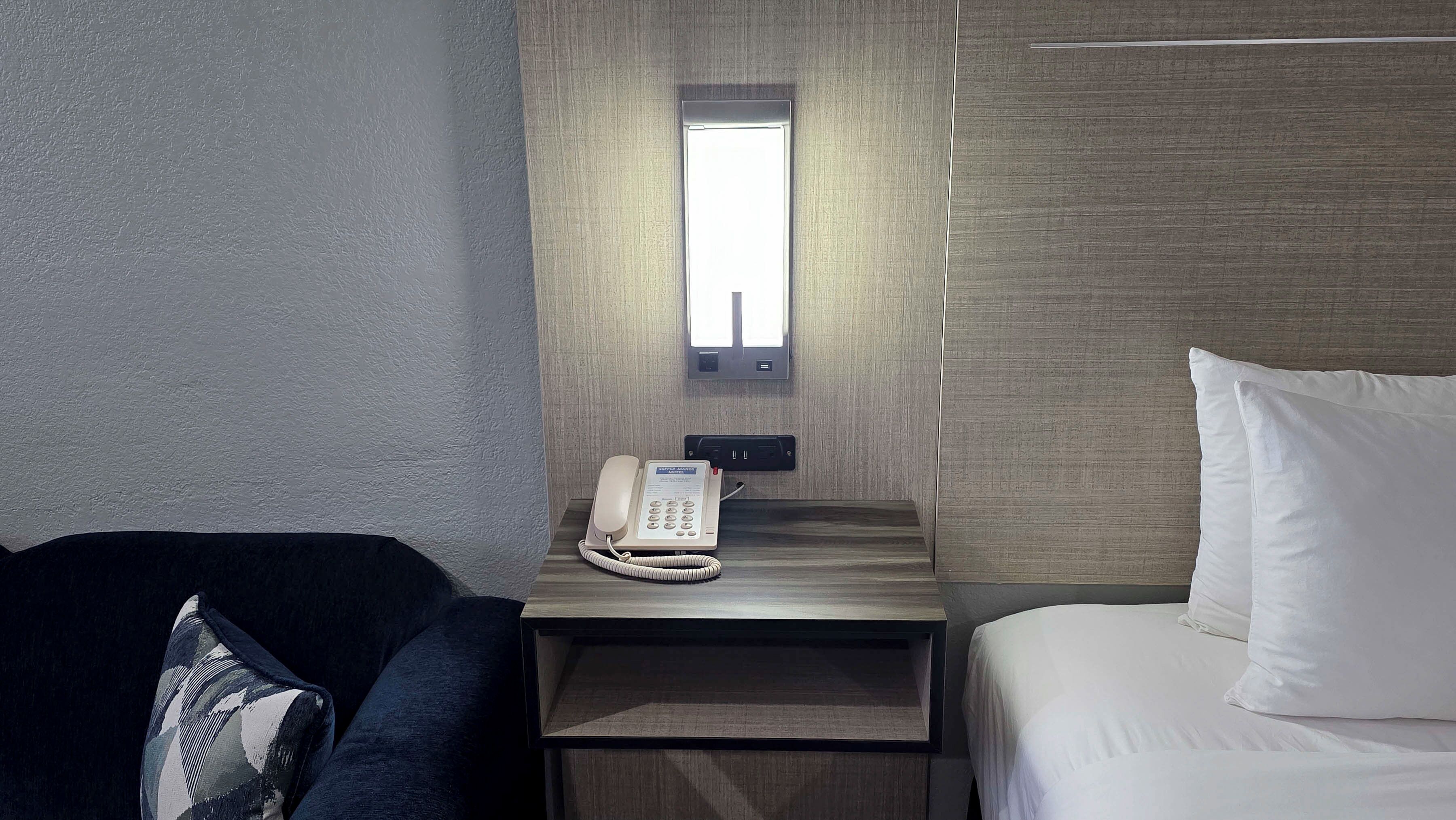 A hotel room with a bed, a bedside table with a phone, a small window, and a wall-mounted light.