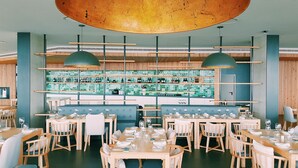 A restaurant with wooden tables and chairs, a bar area with shelves of bottles, and a large circular ceiling light. - Pedras do Mar Resort & SPA (Ponta Delgada)
